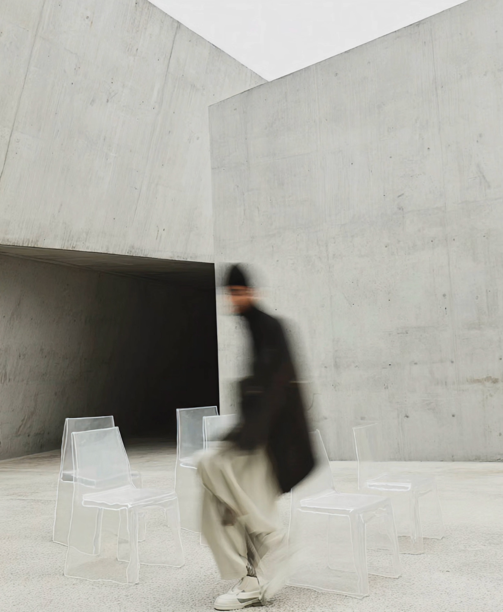 A figure moving through a concrete gallery lined with translucent chairs